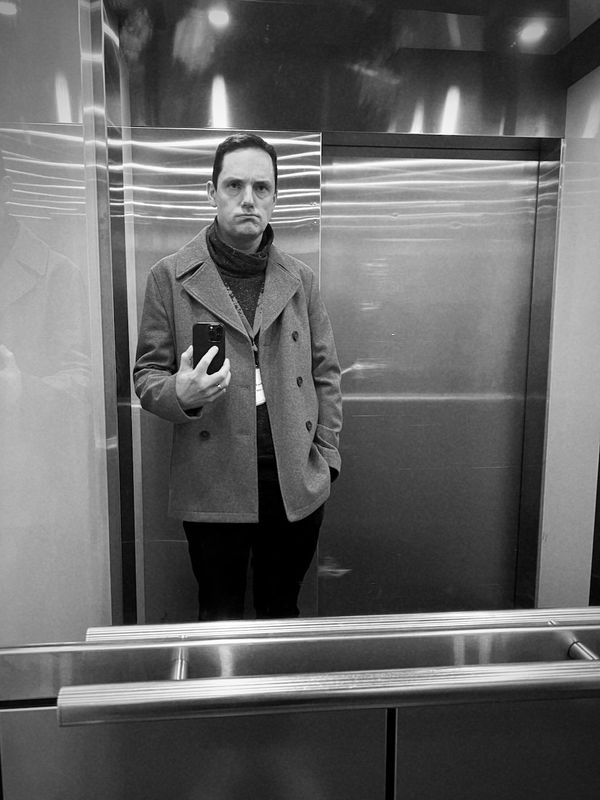 A black and white photo of Adrian Lansdown in a lift, wearing a coat and being grumpy