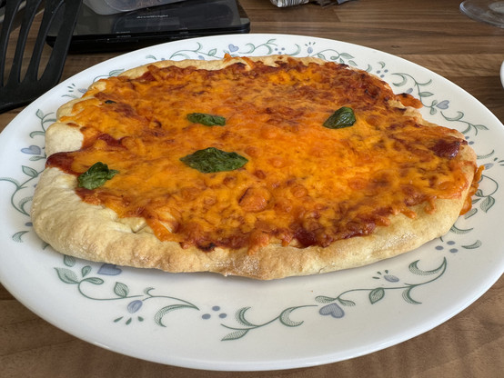 A homemade pizza with a golden brown crust, generously topped with melted cheese and tomato sauce, garnished with basil leaves, served on a white plate with green floral patterns.