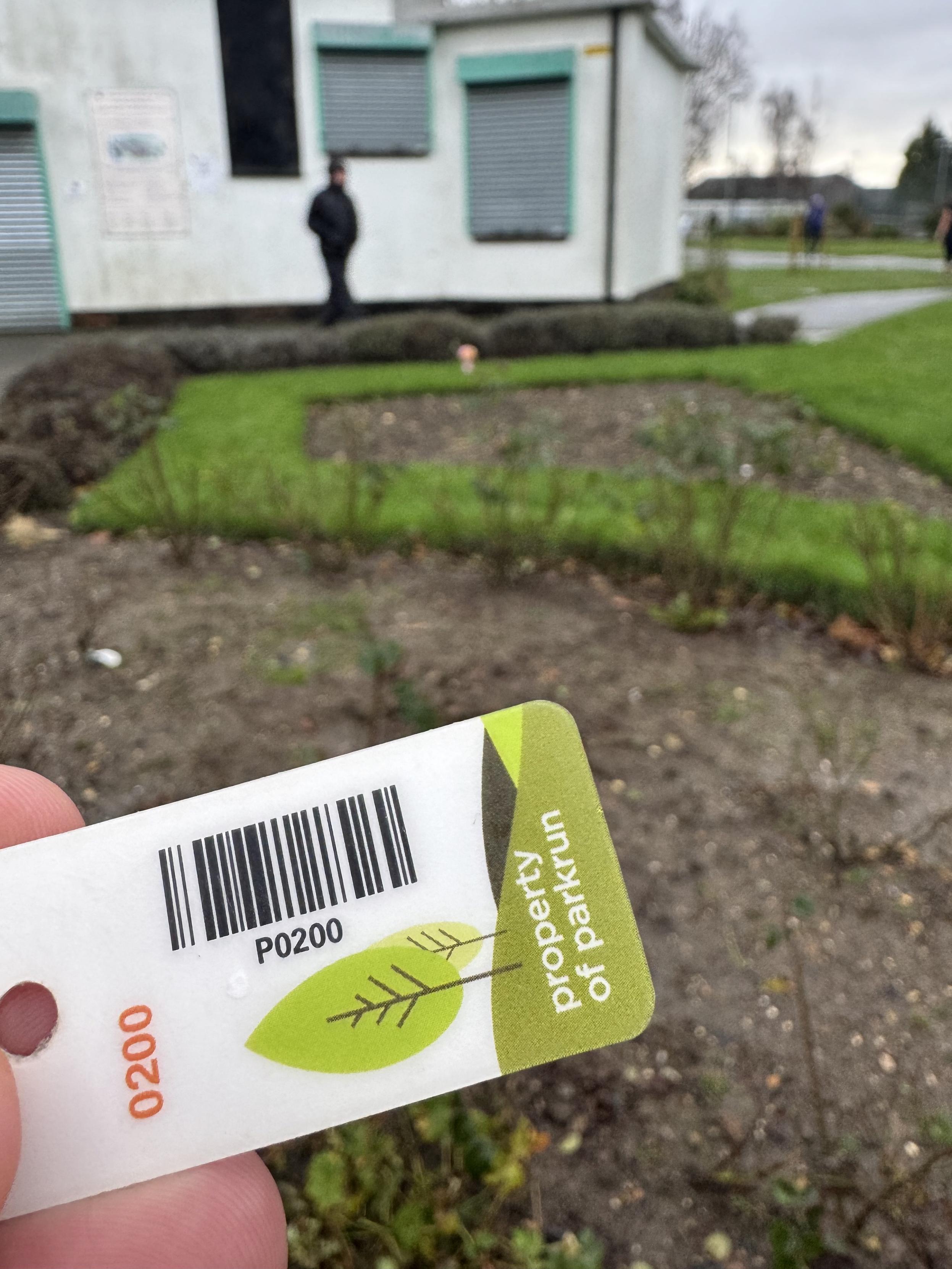 ParkRun token with a barcode and numbers 0200 printed on it.
In front of a mud patch and garden in blurred distance
