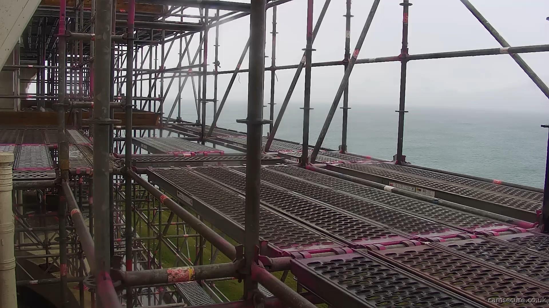 Webcam view of a sea view in the background. In the foreground is some rusty red pipes and metal scaffolding.